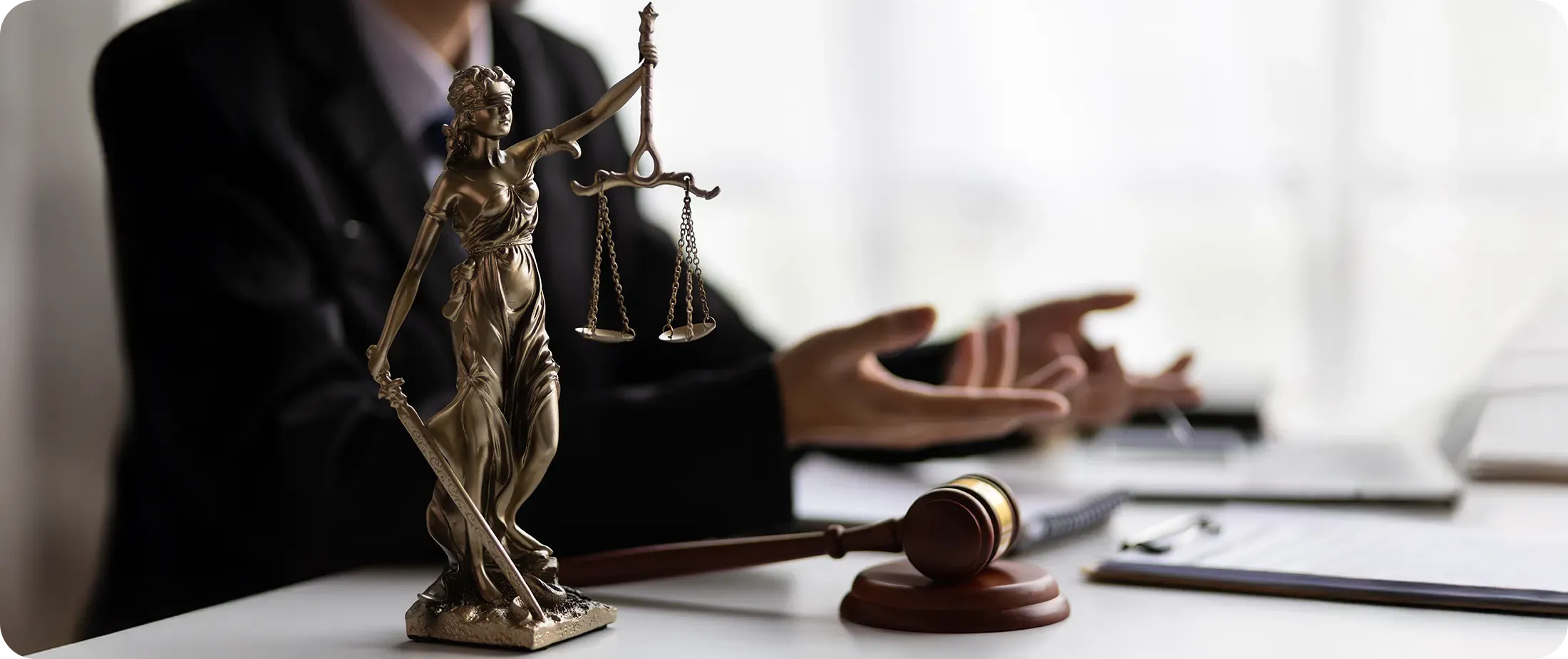 A bronze Lady Justice statue and a wooden gavel on a desk, with a lawyer gesturing in discussion in the background