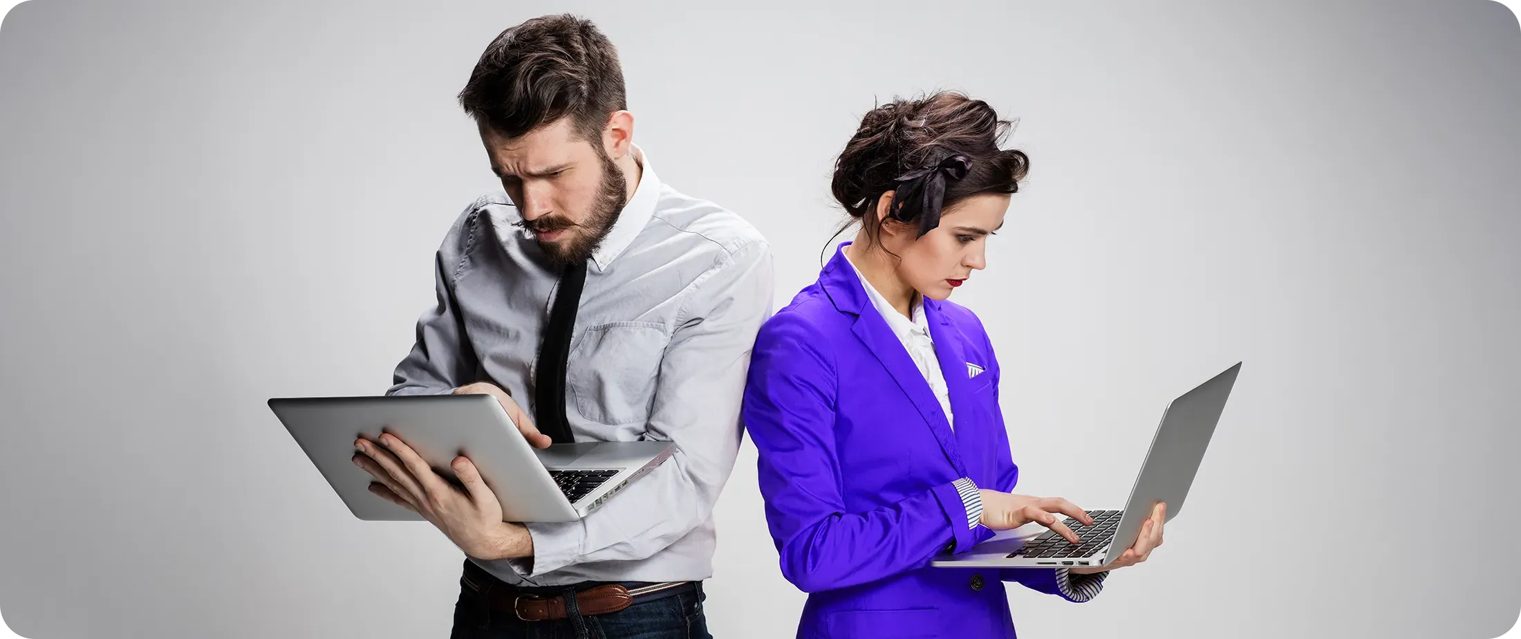 Two professionals standing back to back, each focused on their own laptop, suggesting parallel workflows